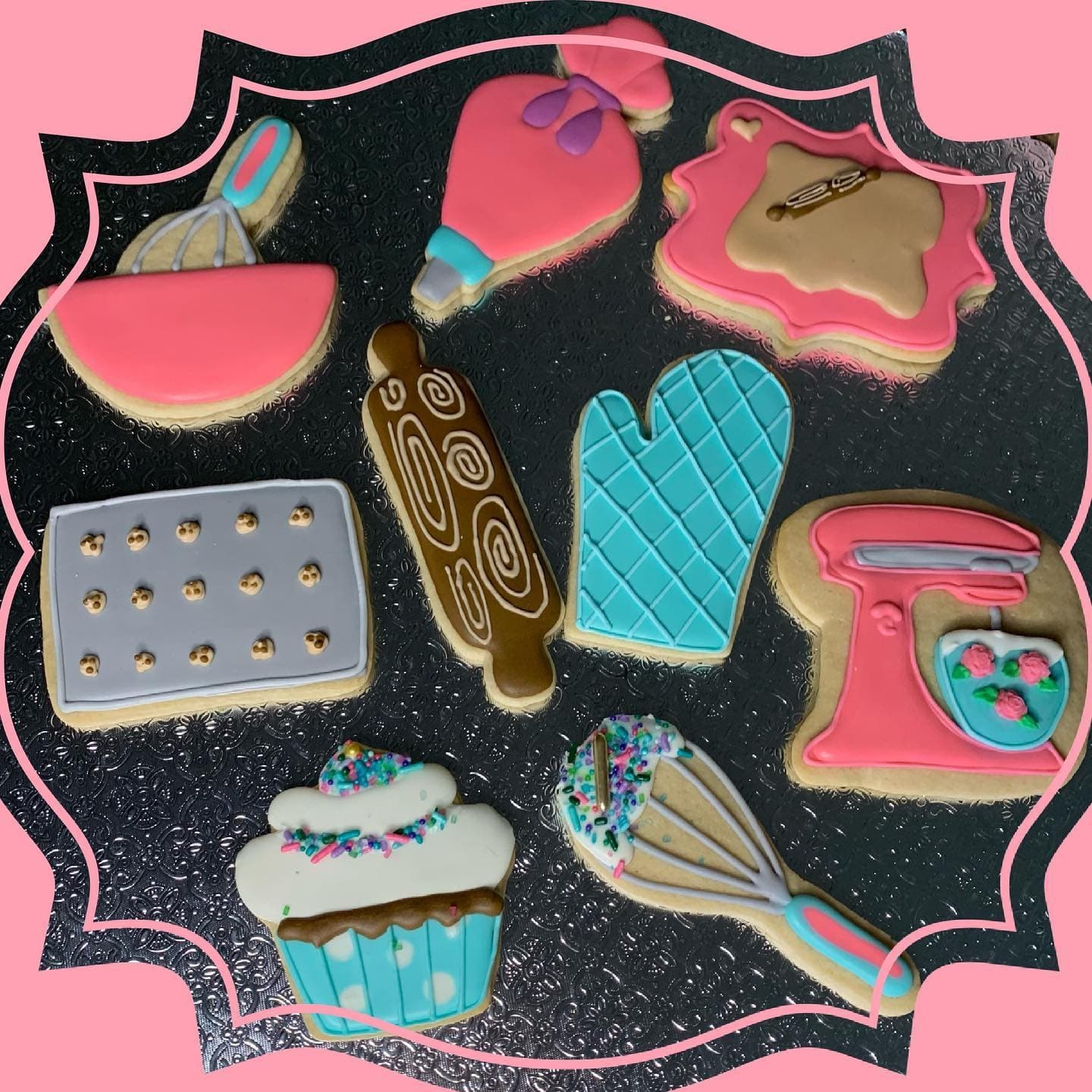 Pink stand mixer, whisk, oven mitt, rolling pin and cupcake cookies on black inside a pink scalloped frame