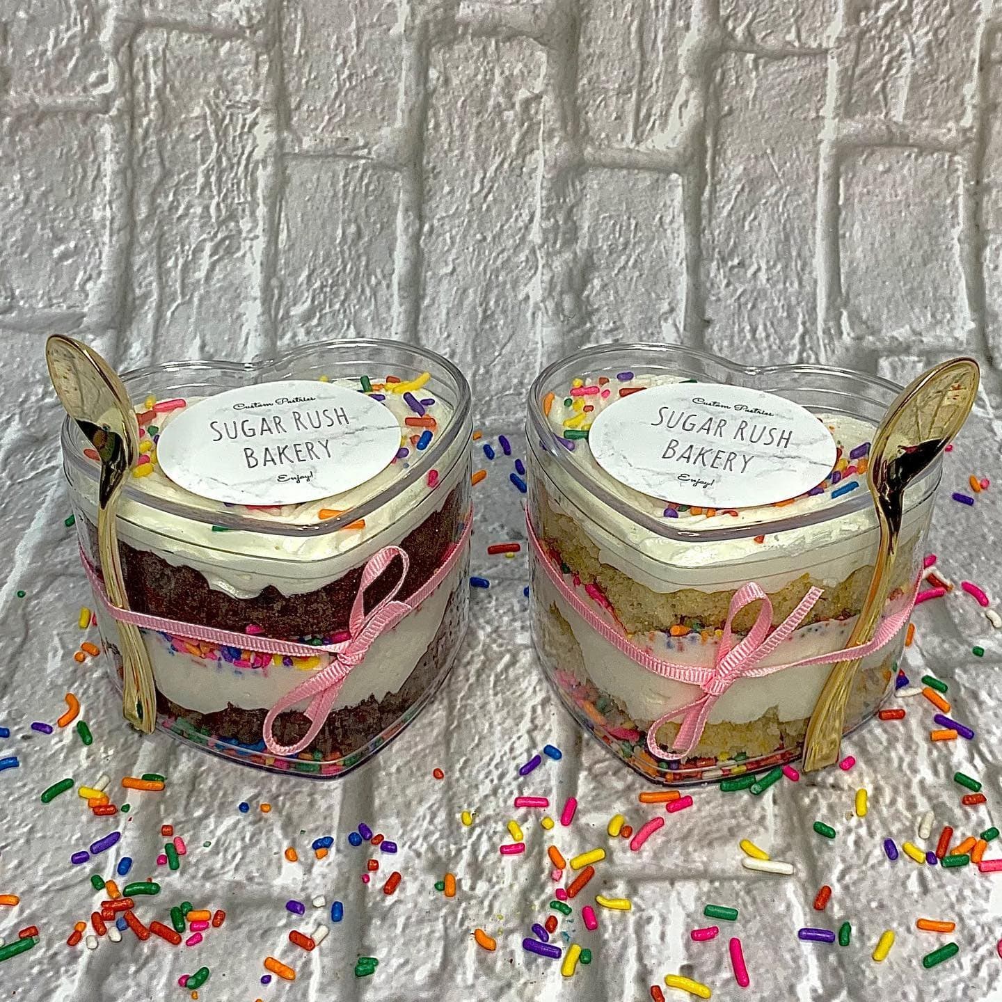 Heart-shaped clear jars layered with cake and frosting, tied with pink ribbon and gold spoons