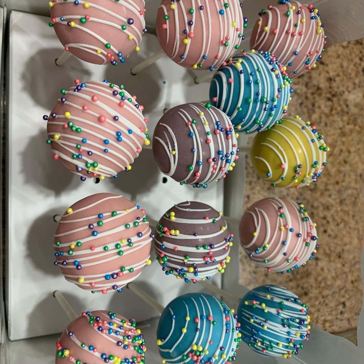Pink, teal and yellow cake pops with drizzle and sprinkles standing in a display box