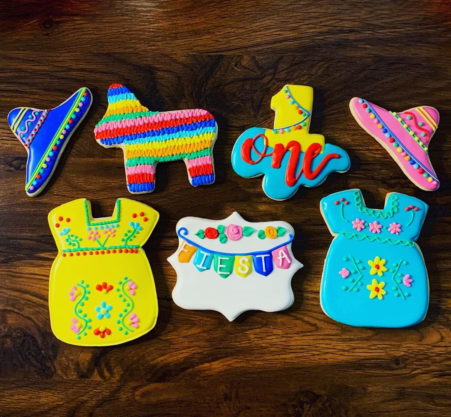 Bright piñata, sombrero and embroidered-dress cookies with a pink one plaque on dark wood