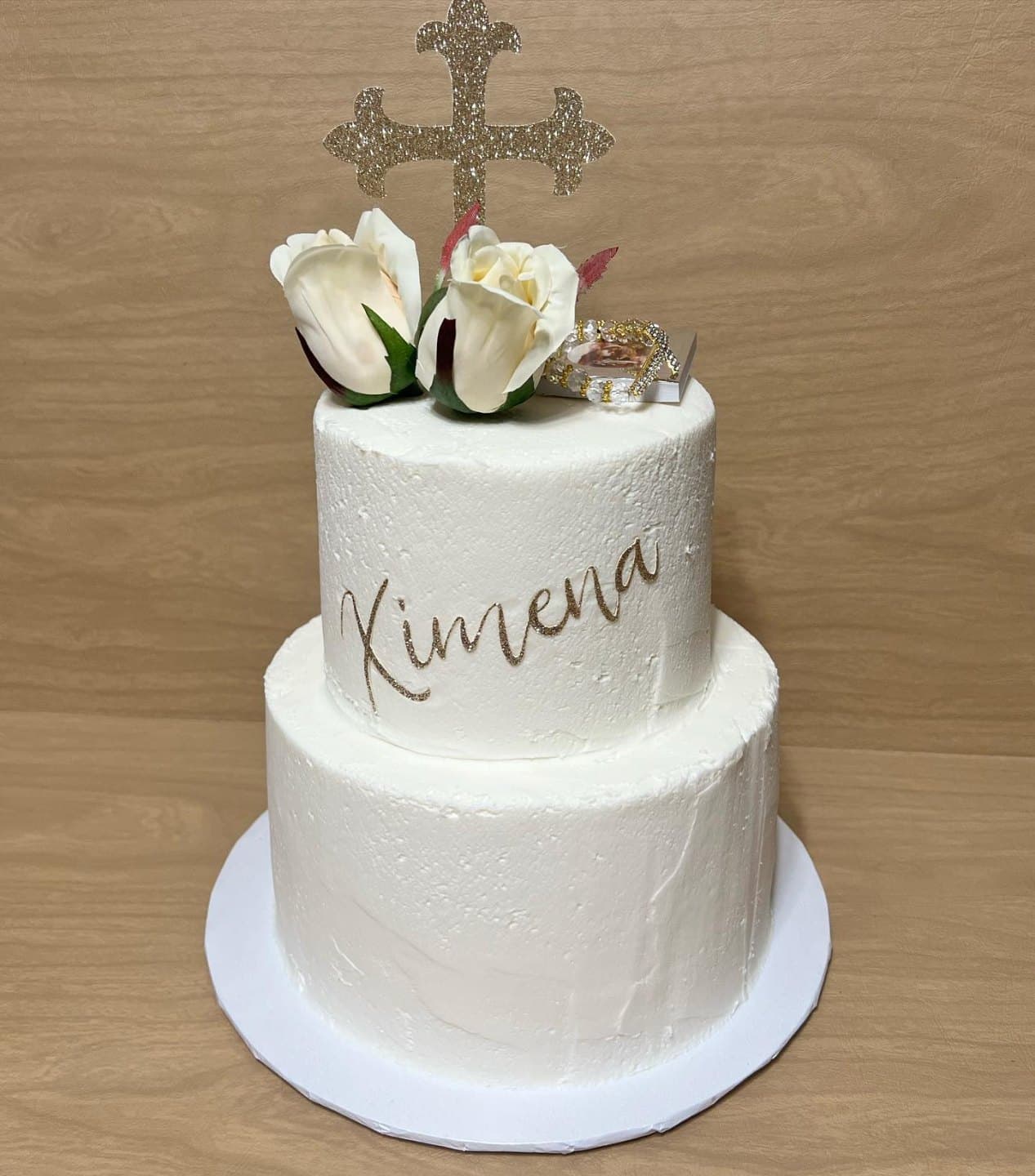 Two-tier textured white buttercream cake with a gold script name, white roses and a glittering cross on top